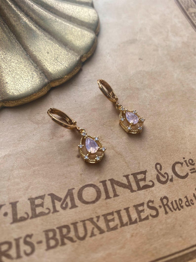 May include: A pair of gold hoop earrings with a teardrop-shaped pink stone surrounded by smaller clear stones.