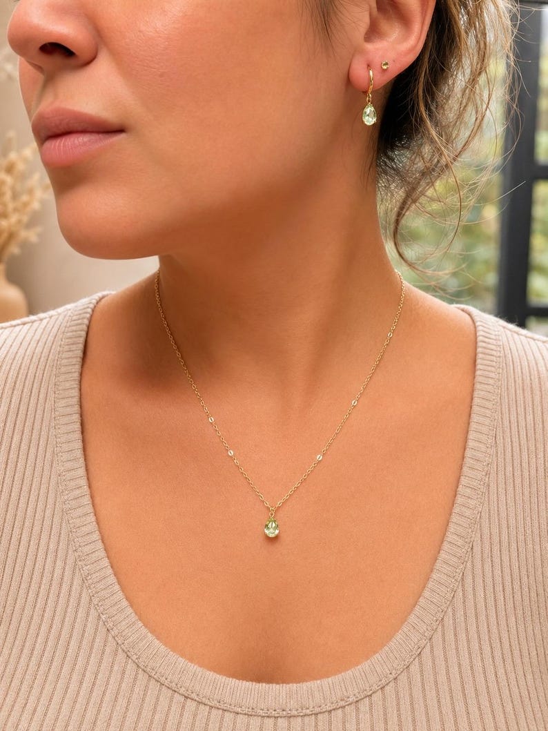 May include: A gold chain necklace featuring a small, light green, teardrop-shaped pendant. The set includes matching gold hoop earrings, each adorned with a single, light green, teardrop-shaped charm that hangs from the bottom of the hoop.