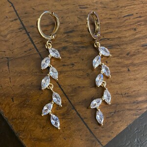 May include: A pair of gold-colored hoop earrings with a dangling leaf design. Each earring features a series of clear, marquise-cut crystals set along a delicate chain, creating a sparkling, elegant look. The earrings are displayed on a wooden surface.