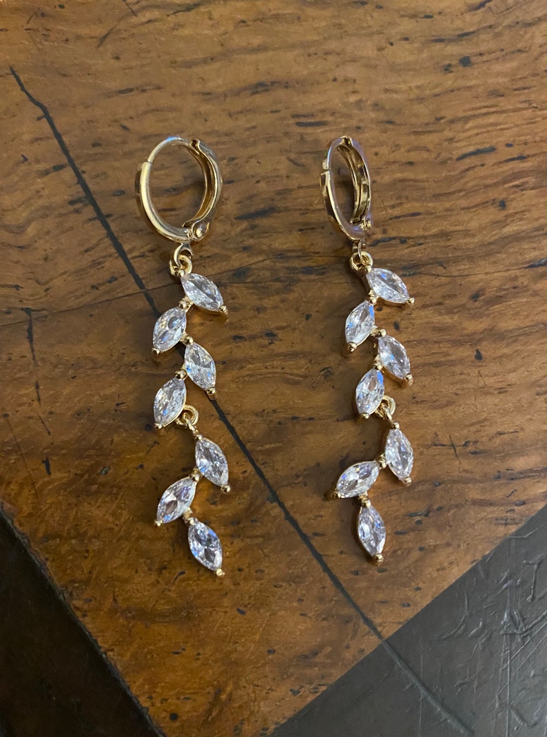 May include: A pair of gold-colored hoop earrings with a dangling leaf design. Each earring features a series of clear, marquise-cut crystals set along a delicate chain, creating a sparkling, elegant look. The earrings are displayed on a wooden surface.