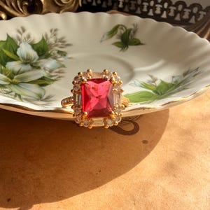 May include: A gold ring featuring a large, rectangular, vibrant red center stone surrounded by a halo of smaller, clear, baguette-cut and round-cut stones. The band is thin and metallic gold, with a textured surface along the outer edges.