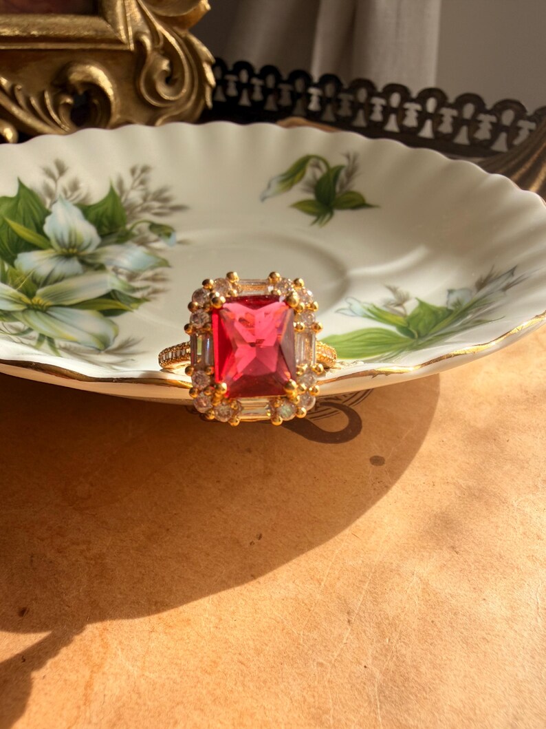 May include: A gold ring featuring a large, rectangular, vibrant red center stone surrounded by a halo of smaller, clear, baguette-cut and round-cut stones. The band is thin and metallic gold, with a textured surface along the outer edges.