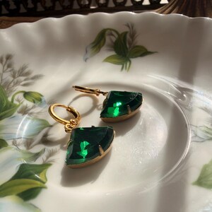May include: A pair of gold-toned hoop earrings featuring faceted, fan-shaped emerald green stones. Each earring has a hinged closure and a polished metal setting that secures the vibrant green gemstone, creating a distinct geometric silhouette.