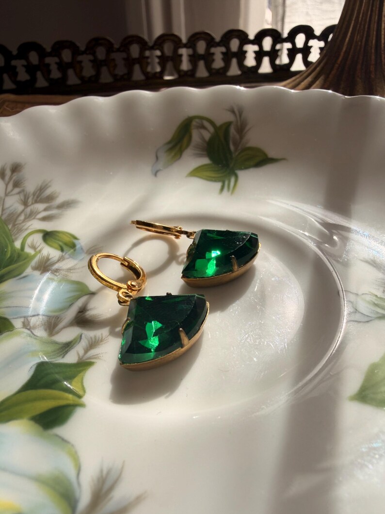 May include: A pair of gold-toned hoop earrings featuring faceted, fan-shaped emerald green stones. Each earring has a hinged closure and a polished metal setting that secures the vibrant green gemstone, creating a distinct geometric silhouette.