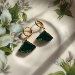 May include: A pair of gold-toned hoop earrings featuring dangling, fan-shaped, deep emerald green faceted gemstones. Each earring is suspended from a small, circular gold-toned hoop closure, showcasing a clear, polished finish on the stones.