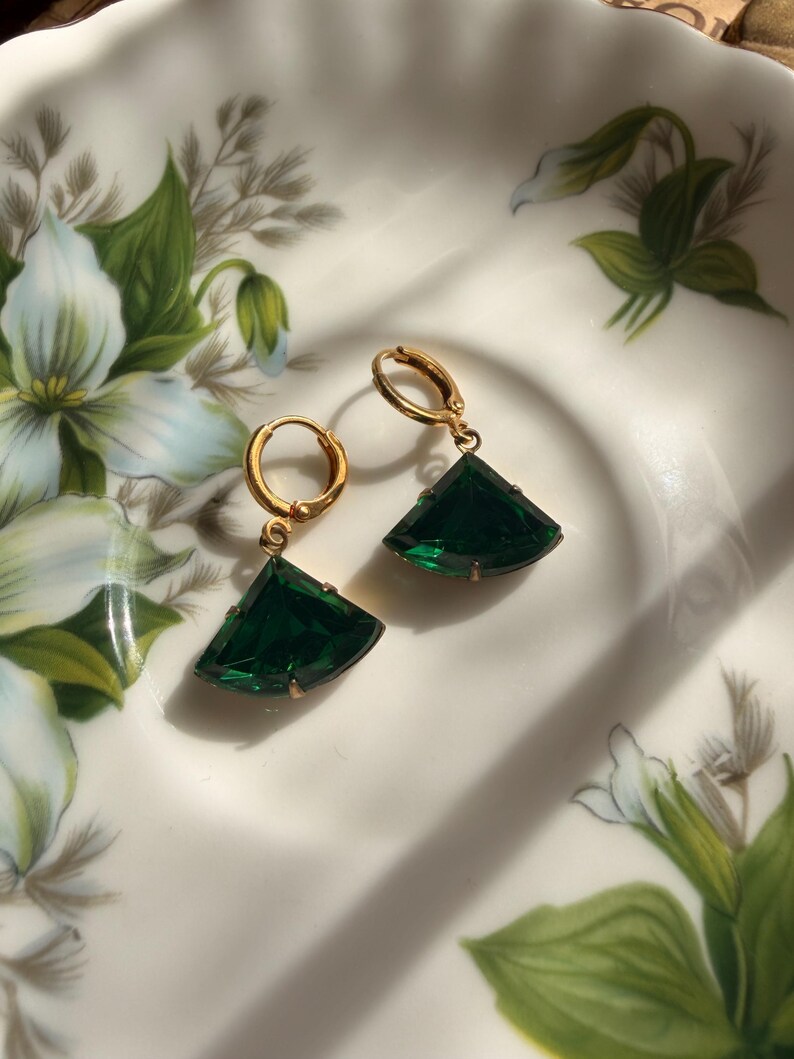 May include: A pair of gold-toned hoop earrings featuring dangling, fan-shaped, deep emerald green faceted gemstones. Each earring is suspended from a small, circular gold-toned hoop closure, showcasing a clear, polished finish on the stones.