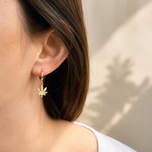 May include: Gold-colored hoop earrings with a cannabis leaf charm. The earrings are small and delicate, with a shiny finish. The cannabis leaf charm is detailed and adds a unique touch to the earrings. The earrings are shown on a person's ear.