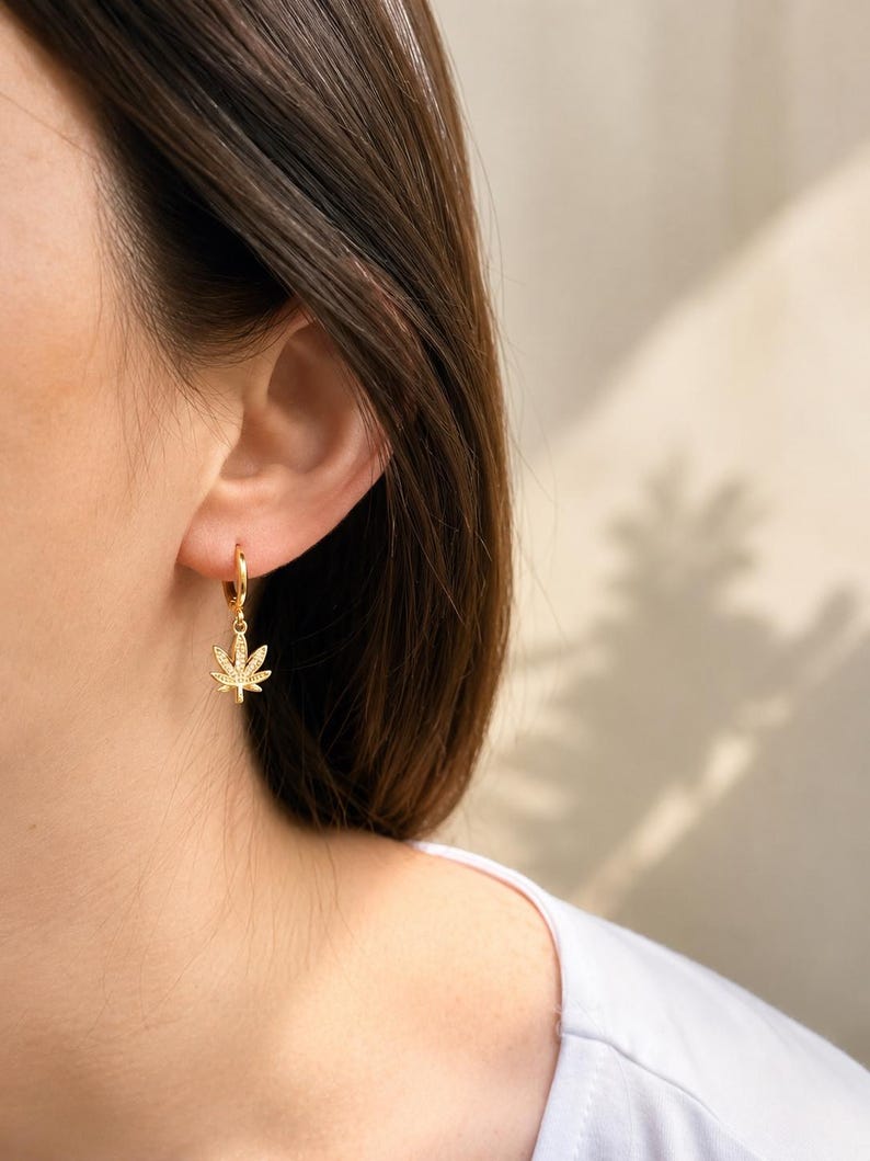 May include: Gold-colored hoop earrings with a cannabis leaf charm. The earrings are small and delicate, with a shiny finish. The cannabis leaf charm is detailed and adds a unique touch to the earrings. The earrings are shown on a person's ear.