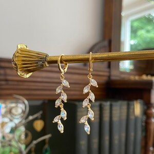 May include: Gold-tone earrings with a leaf design. Each earring features a small hoop and a dangling arrangement of clear, marquise-cut crystals. The earrings are displayed on a gold-colored rod against a blurred background.