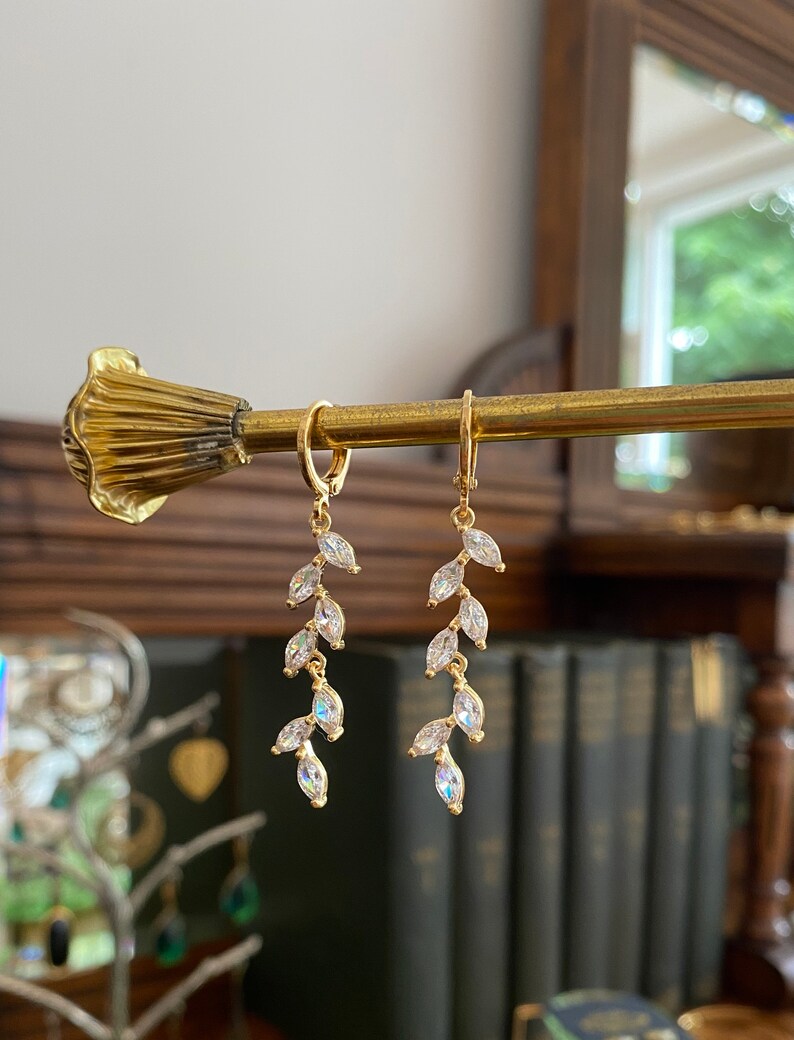 May include: Gold-tone earrings with a leaf design. Each earring features a small hoop and a dangling arrangement of clear, marquise-cut crystals. The earrings are displayed on a gold-colored rod against a blurred background.
