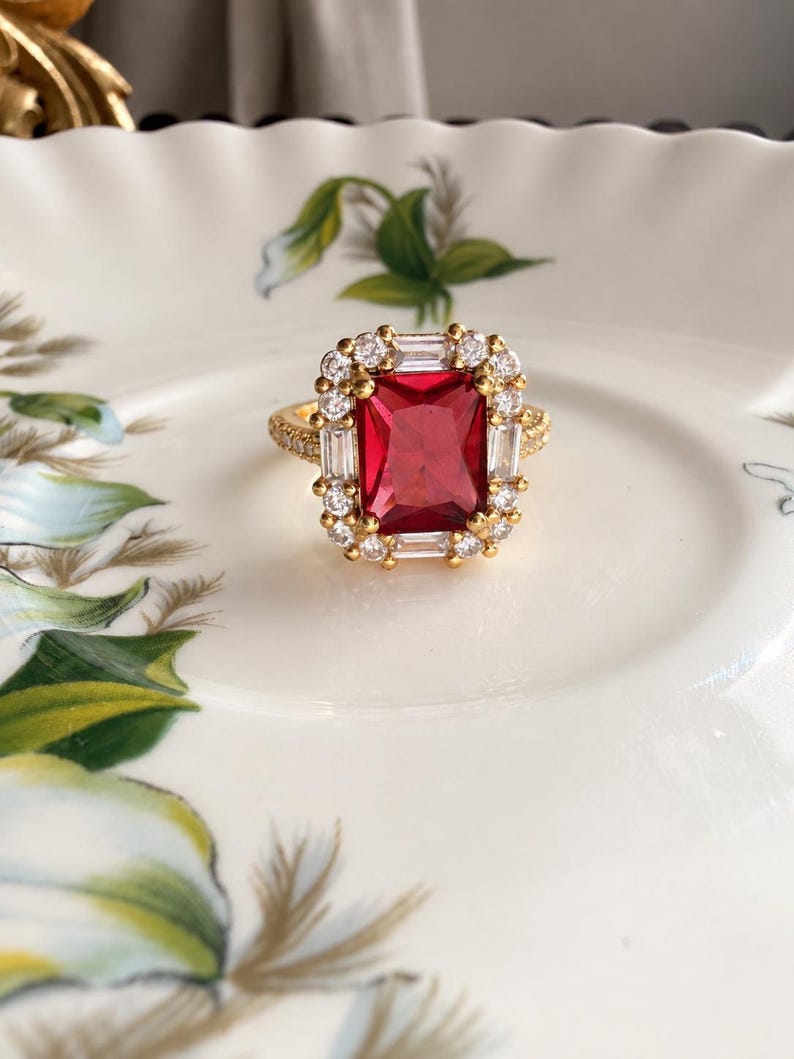 May include: A gold ring featuring a large rectangular red center stone surrounded by a halo of alternating baguette and round clear stones. The band is thin and gold, with small clear stones set along the visible portion of the ring shank.