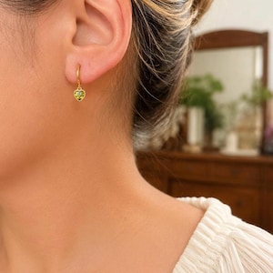 K&ouml;nnte beinhalten: Ein goldener Creolen-Ohrring mit einem kleinen, herzf&ouml;rmigen Anh&auml;nger, der in der Mitte einen facettierten, hellgr&uuml;nen Edelstein tr&auml;gt. Der Ohrring ist am Ohrl&auml;ppchen befestigt und zeichnet sich durch ein poliertes Goldfinish aus.