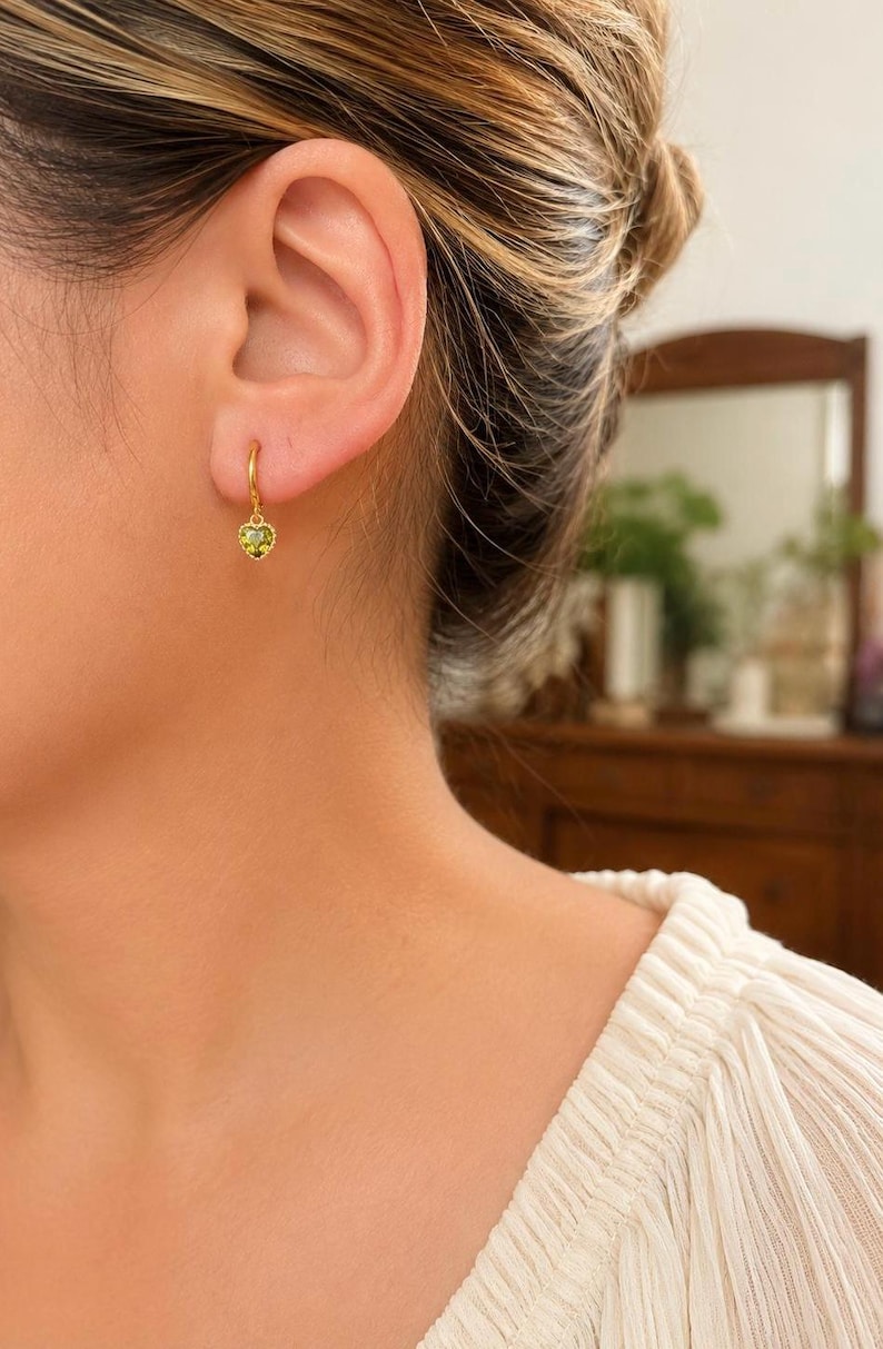 K&ouml;nnte beinhalten: Ein goldener Creolen-Ohrring mit einem kleinen, herzf&ouml;rmigen Anh&auml;nger, der in der Mitte einen facettierten, hellgr&uuml;nen Edelstein tr&auml;gt. Der Ohrring ist am Ohrl&auml;ppchen befestigt und zeichnet sich durch ein poliertes Goldfinish aus.