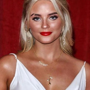 May include: A woman with blonde hair wearing a white sleeveless dress and a gold necklace with a rose pendant. She is wearing red lipstick and has a red background behind her.