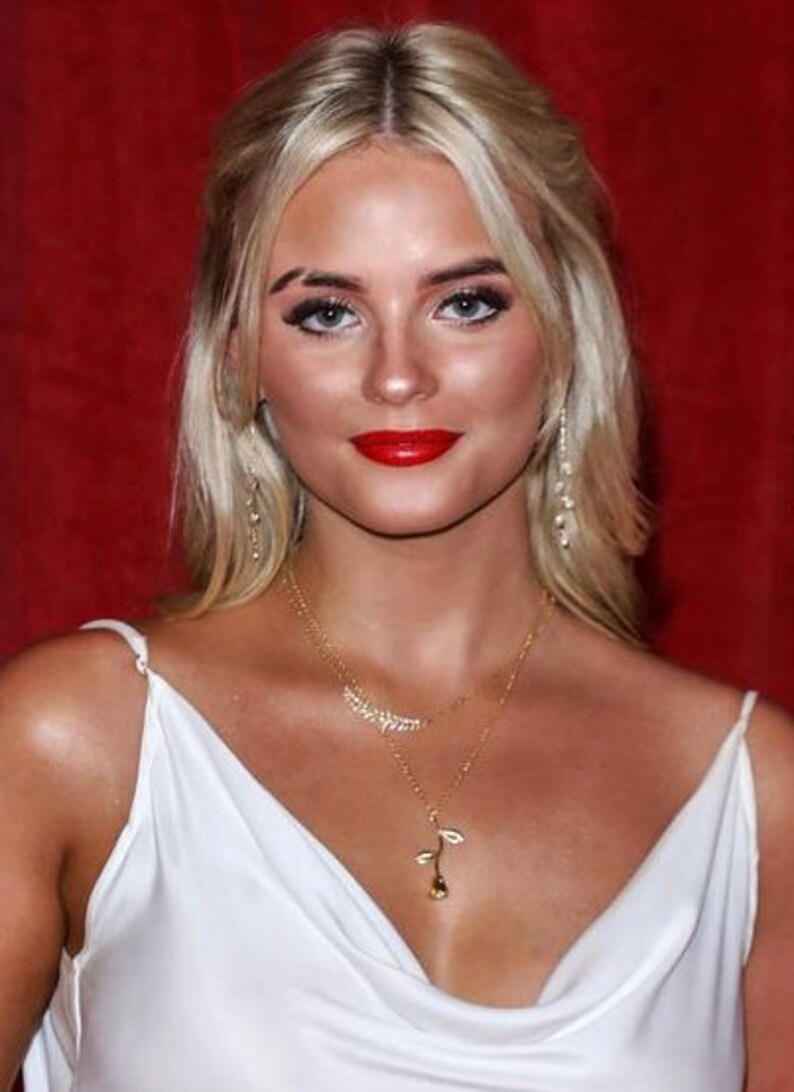 May include: A woman with blonde hair wearing a white sleeveless dress and a gold necklace with a rose pendant. She is wearing red lipstick and has a red background behind her.