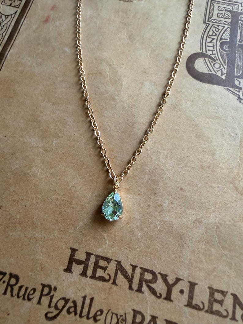 May include: A gold-toned chain necklace featuring a single pear-shaped, light green gemstone pendant. The pendant is secured in a simple metal setting, and the delicate chain consists of small, uniform oval links that connect to the top of the stone.