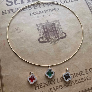 Pode incluir: Um colar de corrente de ouro com três pingentes em forma de trevo. Os pingentes são adornados com strass transparentes e apresentam pedras preciosas vermelhas, verdes e pretas no centro.