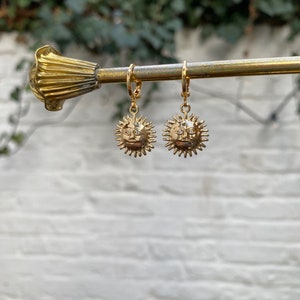 Op de afbeelding: Een paar gouden creolen met een zonnenhanger. De zonnenhanger heeft een gezicht en stralen.