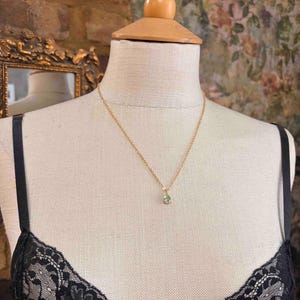 May include: A delicate gold chain necklace featuring a small, teardrop-shaped pendant with a pale green gemstone. The necklace is displayed on a beige fabric mannequin wearing a black lace garment with thin straps, showcasing the jewelry piece.