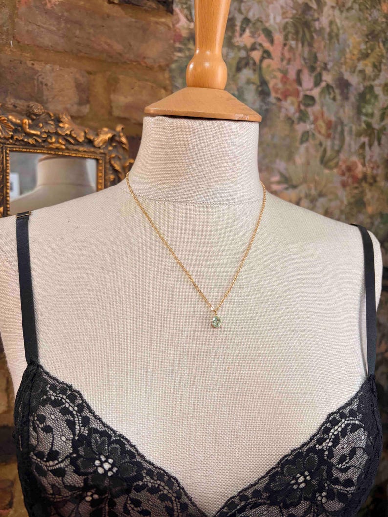 May include: A delicate gold chain necklace featuring a small, teardrop-shaped pendant with a pale green gemstone. The necklace is displayed on a beige fabric mannequin wearing a black lace garment with thin straps, showcasing the jewelry piece.