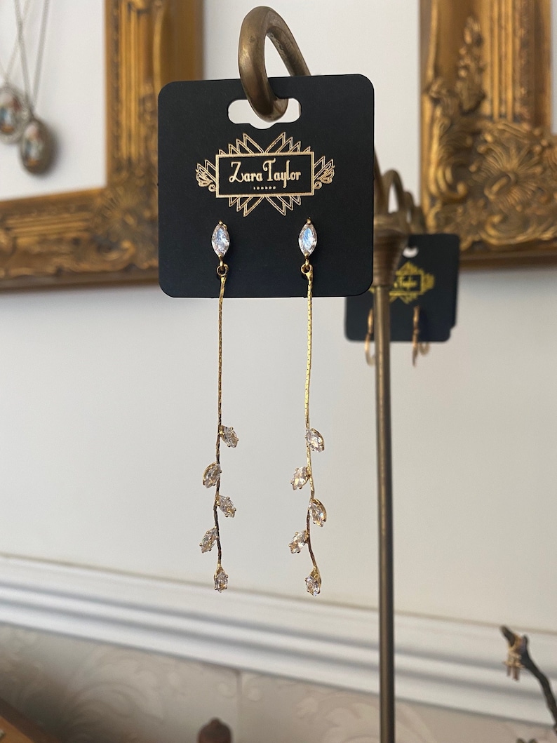 May include: A pair of gold dangle earrings with a delicate leaf design and clear crystal accents. The earrings are displayed on a black card with the text "Zara Taylor" printed on it.