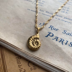Puede incluir: Un collar de cadena dorada con un colgante de concha de nautilus dorada. El collar está sobre un libro antiguo con texto en francés.