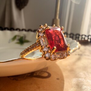May include: A gold ring featuring a large rectangular red center stone surrounded by a halo of smaller clear stones. The band is thin and encrusted with tiny clear stones along the sides, creating a bright, reflective appearance in the sunlight.