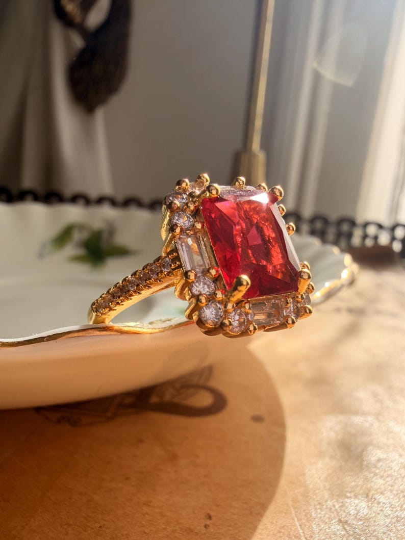 May include: A gold ring featuring a large rectangular red center stone surrounded by a halo of smaller clear stones. The band is thin and encrusted with tiny clear stones along the sides, creating a bright, reflective appearance in the sunlight.