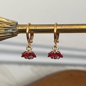 Peut inclure: Une paire de boucles d'oreilles créoles dorées avec de petites breloques en forme de fleur. Chaque breloque présente un design de pétale avec une pierre rouge centrale, suspendue à un anneau doré simple doté d'un fermoir à charnière.