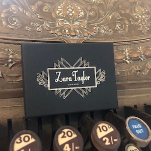May include: A black box with the text "Zara Taylor London" in a white, decorative font. The box is resting on the keys of an antique cash register. The cash register keys have numbers and prices.