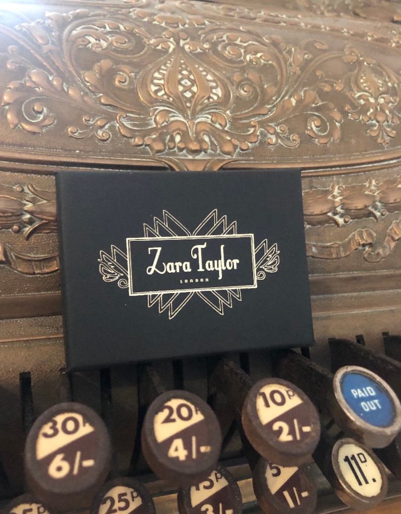 May include: A black box with the text "Zara Taylor London" in a white, decorative font. The box is resting on the keys of an antique cash register. The cash register keys have numbers and prices.