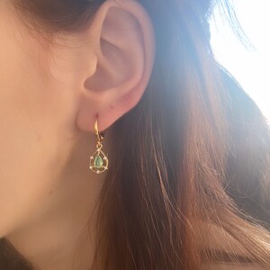 May include: Gold hoop earrings with a teardrop-shaped green gemstone and small clear stones around the gemstone.
