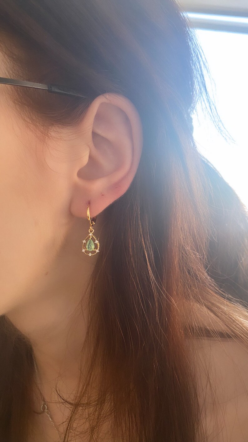 May include: Gold hoop earrings with a teardrop-shaped green gemstone and small clear stones around the gemstone.
