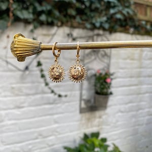 Op de afbeelding: Een paar gouden creolen met een zon hanger. De zon hanger heeft een gezicht en stralen.