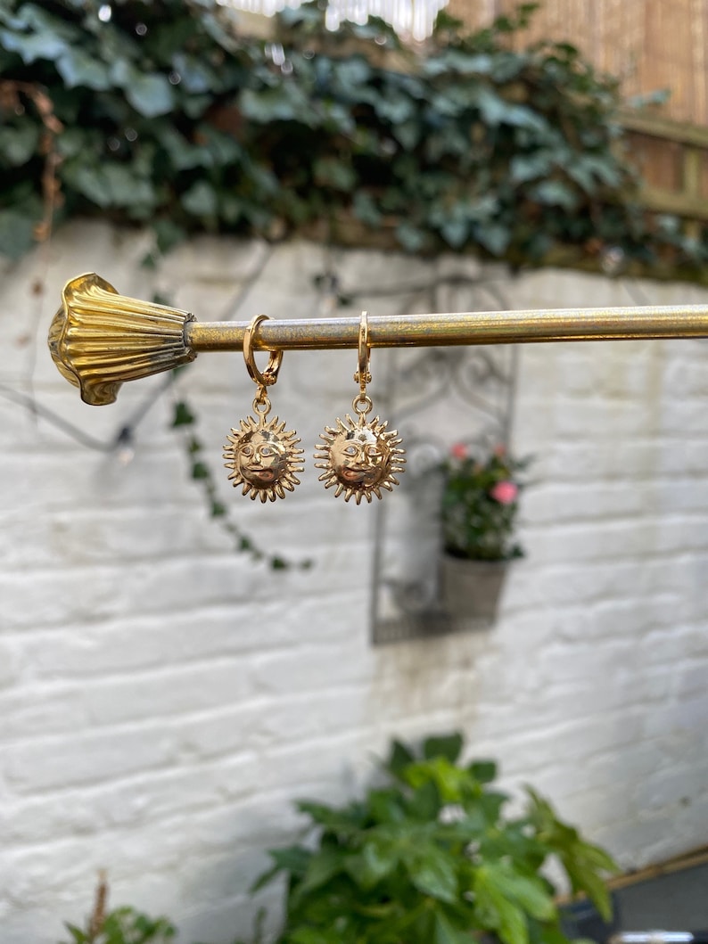 Op de afbeelding: Een paar gouden creolen met een zon hanger. De zon hanger heeft een gezicht en stralen.
