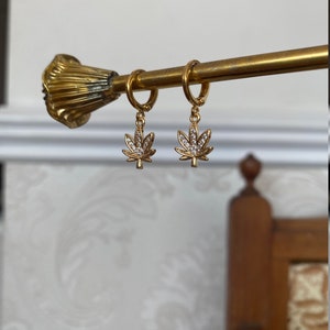 May include: Gold-colored hoop earrings with a cannabis leaf charm. Each charm is a detailed leaf shape, encrusted with small, clear crystals. The earrings are suspended from a gold-toned bar.