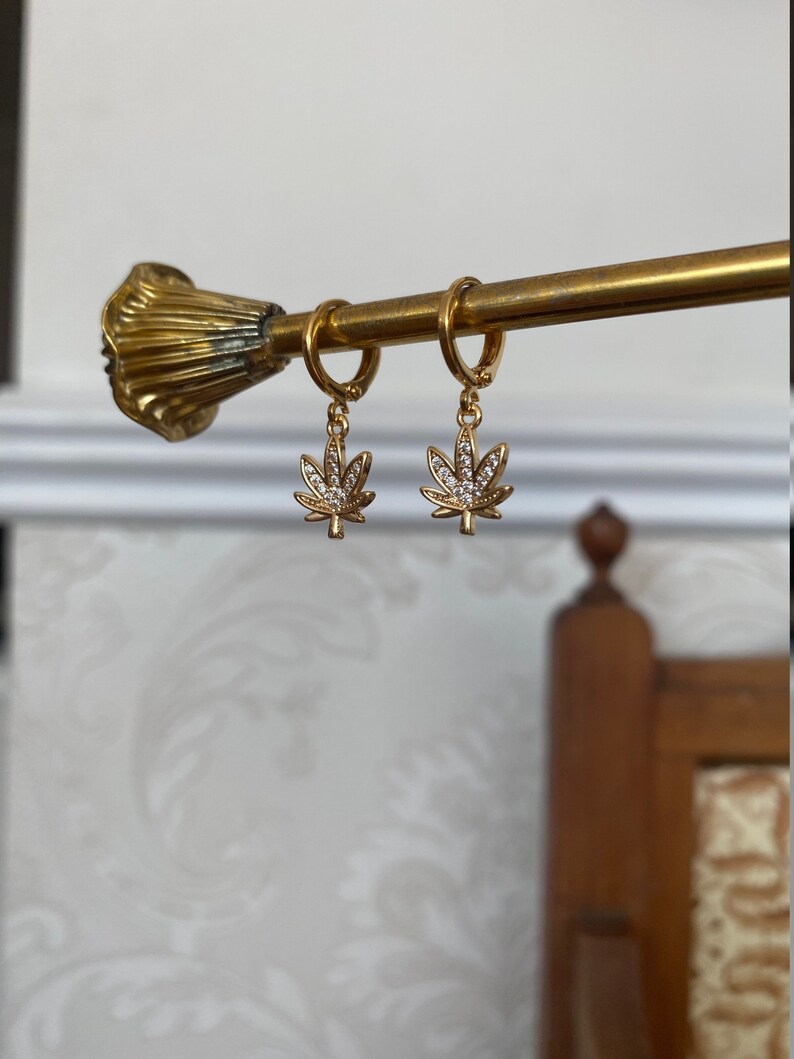 May include: Gold-colored hoop earrings with a cannabis leaf charm. Each charm is a detailed leaf shape, encrusted with small, clear crystals. The earrings are suspended from a gold-toned bar.