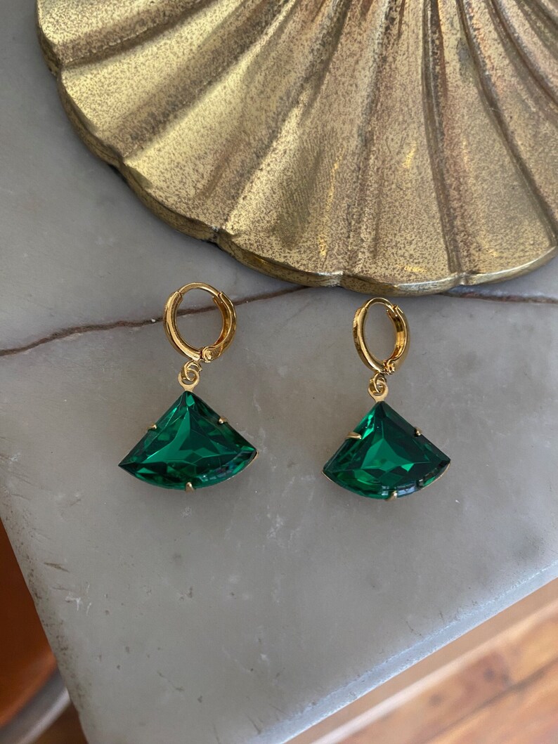 May include: A pair of gold-toned hoop earrings featuring faceted, fan-shaped emerald green glass pendants. Each earring is suspended from a circular gold-toned hoop closure, showcasing a clear, deep green gemstone cut into a wide, geometric fan shape.