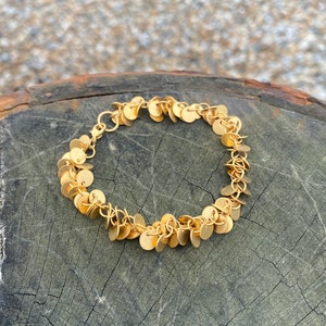 Peut inclure: Un bracelet doré orné de petits disques plats et ronds. Le bracelet est composé de maillons de chaîne délicats et est présenté sur une surface en bois grise et vieillie. Le design du bracelet est simple et élégant.