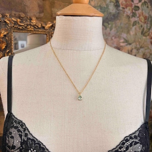 May include: A delicate gold chain necklace featuring a single, small, pear-shaped light green gemstone pendant. The necklace is displayed on a beige fabric mannequin wearing a black lace garment, highlighting the simple and refined design of the jewelry.