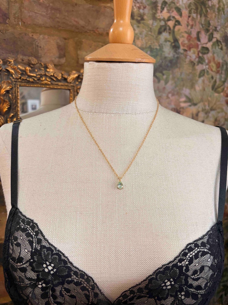 May include: A delicate gold chain necklace featuring a single, small, pear-shaped light green gemstone pendant. The necklace is displayed on a beige fabric mannequin wearing a black lace garment, highlighting the simple and refined design of the jewelry.