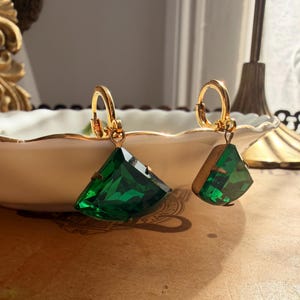 May include: A pair of gold-toned hoop earrings featuring dangling, faceted, fan-shaped emerald green stones. Each earring is suspended from a small, circular gold-colored hoop with a hinged clasp mechanism, showcasing a vibrant, deep green hue.