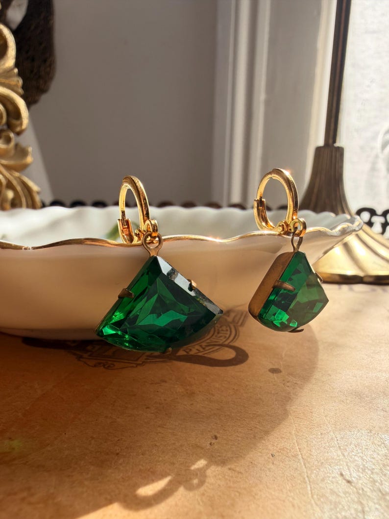 May include: A pair of gold-toned hoop earrings featuring dangling, faceted, fan-shaped emerald green stones. Each earring is suspended from a small, circular gold-colored hoop with a hinged clasp mechanism, showcasing a vibrant, deep green hue.