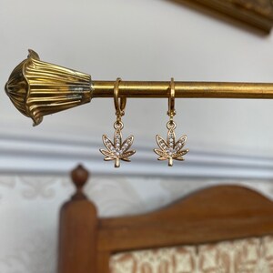 May include: Gold-colored earrings with a cannabis leaf design. Each leaf is adorned with small, clear crystals. The earrings are hanging on a gold-colored rod, showcasing their design and sparkle.
