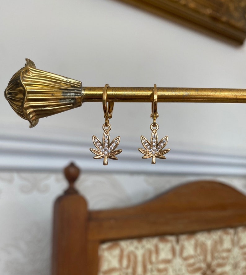 May include: Gold-colored earrings with a cannabis leaf design. Each leaf is adorned with small, clear crystals. The earrings are hanging on a gold-colored rod, showcasing their design and sparkle.