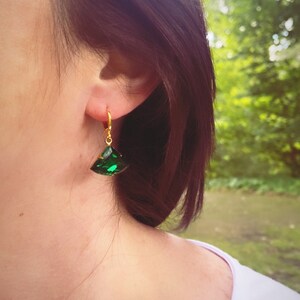 May include: A gold-toned hoop earring featuring a faceted, triangular-shaped emerald green gemstone pendant. The earring is secured with a small hinged clasp mechanism, showcasing a vibrant green hue and a polished metallic gold finish.