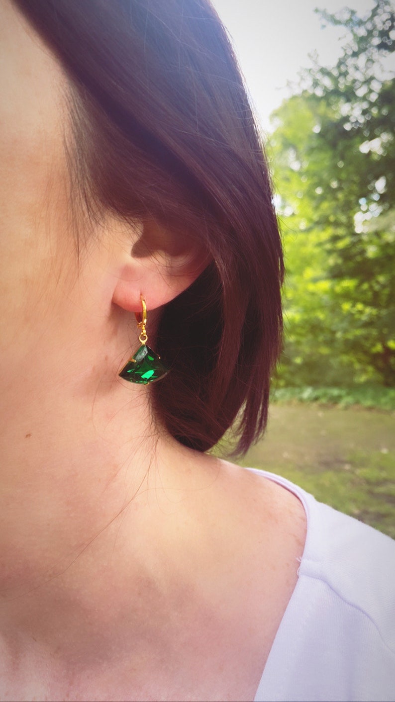 May include: A gold-toned hoop earring featuring a faceted, triangular-shaped emerald green gemstone pendant. The earring is secured with a small hinged clasp mechanism, showcasing a vibrant green hue and a polished metallic gold finish.