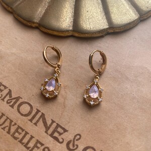 May include: A pair of gold hoop earrings with a teardrop-shaped pendant. Each pendant features a clear pink stone surrounded by smaller clear stones.