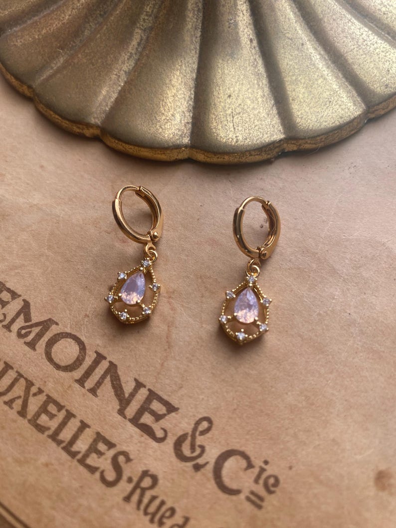 May include: A pair of gold hoop earrings with a teardrop-shaped pendant. Each pendant features a clear pink stone surrounded by smaller clear stones.