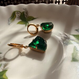 May include: A pair of gold-tone hoop earrings featuring faceted, emerald-green, trapezoidal-shaped stones set in metal prongs. The earrings are designed with a hinged closure mechanism, showcasing a vibrant green hue and a polished metallic finish.